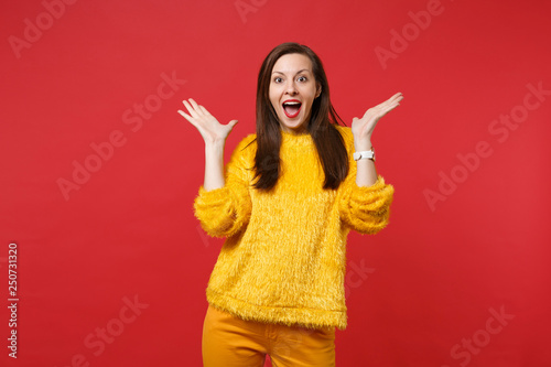 Excited young woman in yellow fur sweater keeping mouth wide open, spreading hands isolated on bright red wall background in studio. People sincere emotions, lifestyle concept. Mock up copy space.