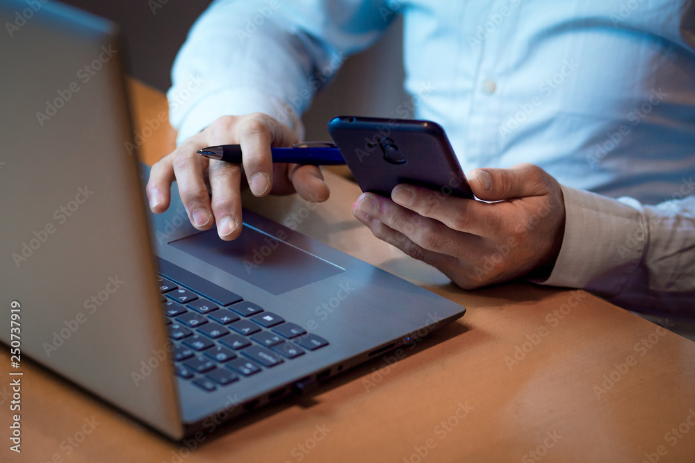 Business concept - Manager's hands holding a smartphone and pen, checking some reports on his laptop Business concept - Manager's hands holding a smartphone and pen, checking some reports on his laptop