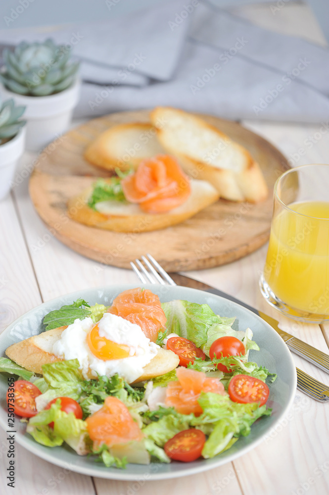 Green salad with lightly salted salmon. On plate with salad toast with poached egg. In the frame of a glass with juice and a plate of bruscheta. Light background. Close-up. Vertical frame orientation.