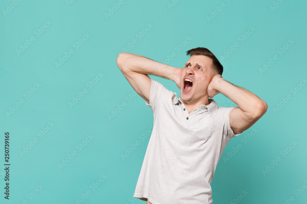 Shocked screaming young man in casual clothes looking up aside covering ears with hands isolated on blue turquoise background in studio. People sincere emotions, lifestyle concept. Mock up copy space. Shocked screaming young man in casual clothes looking up aside covering ears with hands isolated on blue turquoise background in studio. People sincere emotions, lifestyle concept. Mock up copy space.