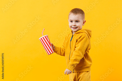 Little fun cheerful kid boy 3-4 years old wearing yellow clothes hold cup of soda isolated on orange wall background, children studio portrait. People, childhood lifestyle concept. Mock up copy space.