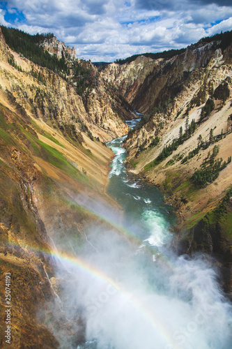 Waterfall with douple rainbow in Yellowstone national park  United States of America  beautiful river in canyon with yellow hills and forest on the top  blue sky with white clounds.  Mountains view. 