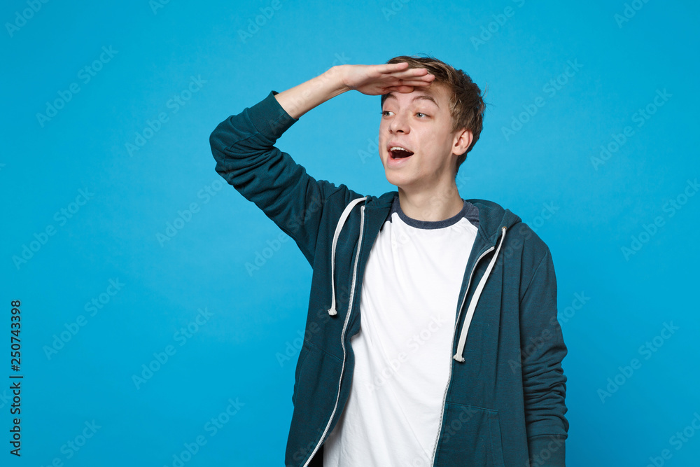 Portrait of curious young man in casual clothes holding hand at forehead looking far away distance isolated on blue background in studio. People sincere emotions lifestyle concept. Mock up copy space. Portrait of curious young man in casual clothes holding hand at forehead looking far away distance isolated on blue background in studio. People sincere emotions lifestyle concept. Mock up copy space.