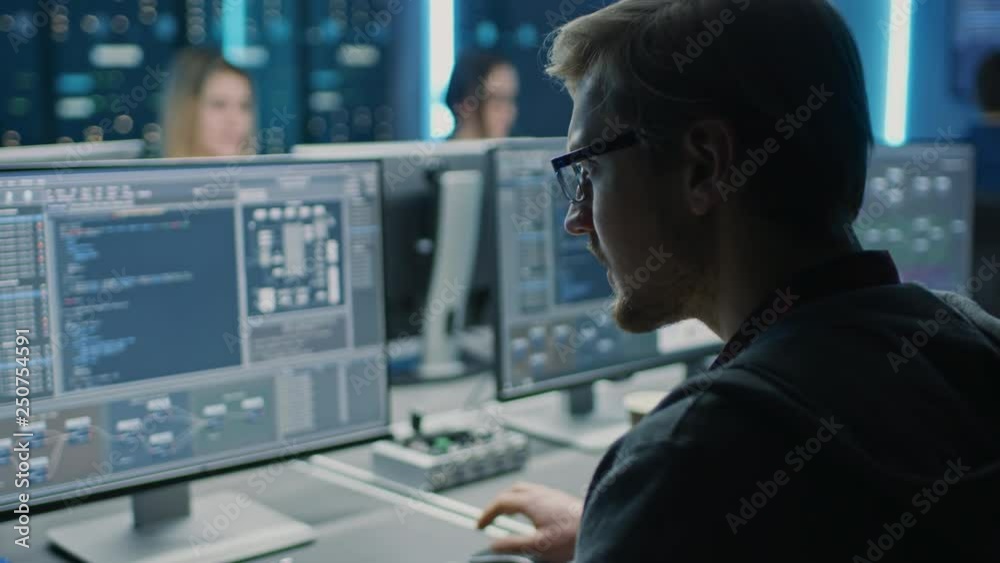 Smart Male IT Programer Working on Desktop Computer in Data Center Technical System Control Room. Team of Young Professionals Programming in Coding Language Smart Male IT Programer Working on Desktop Computer in Data Center Technical System Control Room. Team of Young Professionals Programming in Coding Language