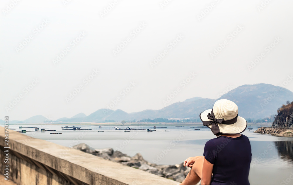 Woman sitting on concrete slab Background The raft floating fish farming in water and mountains at Krasiew dam , Supanburi Thailand