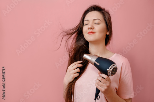 young woman makes hair volume with hairdryer in hand on pink background