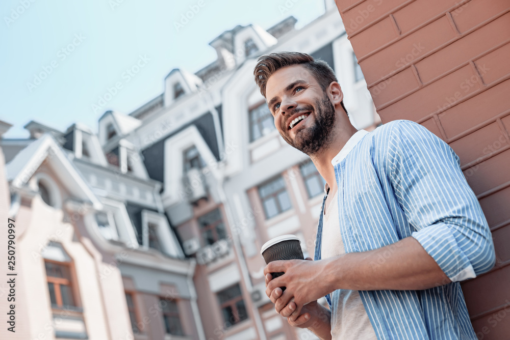 Fototapeta premium Success is peace of mind. Portrait of confident brown-haired man holding coffee cup and smiling aside while walking outdoors