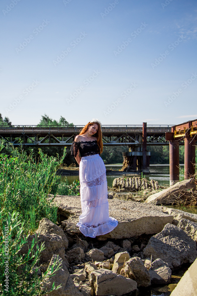 Young redhead woman in dress on the rock near the bridge