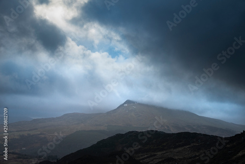 Moody and dramatic Winter landscape image of Moel Saibod from Crimpiau in Snowdonia with stunning shafts of light in stormy weather