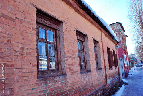 Old red brick building wall with windows close up, small town winter street, snow on roof and poplar tree without leaves, bright blue sky