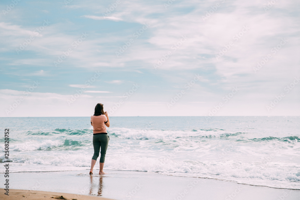 Young Caucasian Woman Standing On Summer Sea Ocean Beach And Taking Photos On Smartphone.