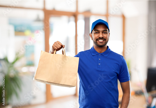 takeaway service and people concept - happy indian delivery man food in paper bag in blue uniform over office background