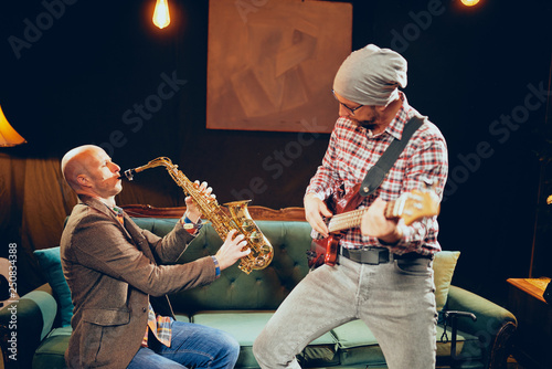 Young Caucasian man with hat on head playing bass guitar while standing in home studio. Next to hix saxophonist. photo