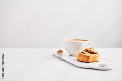 Freshly baked cinnamon roll with spices and cocoa filling and coffee or cappuccino on white serving plate on white marble table over white background. Swedish breakfast. Copy space. Selective focus.