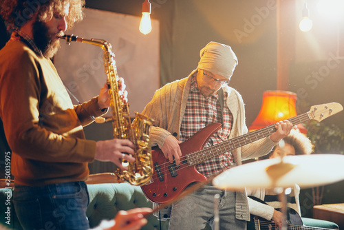 Young Caucasian man with hat on head playing bass guitar while sitting in home studio. In background saxophonist playing his instrument. photo