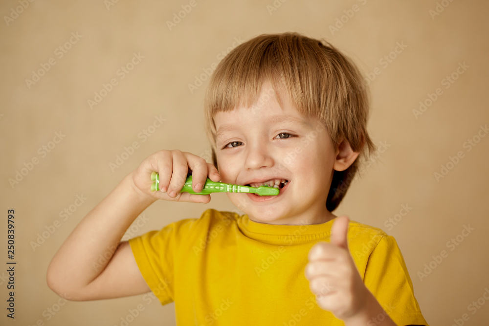 Cute little blond boy 4-5 years old in a yellow T-shirt brushes his teeth with a colorful baby toothbrush looking at the camera Cute little blond boy 4-5 years old in a yellow T-shirt brushes his teeth with a colorful baby toothbrush looking at the camera