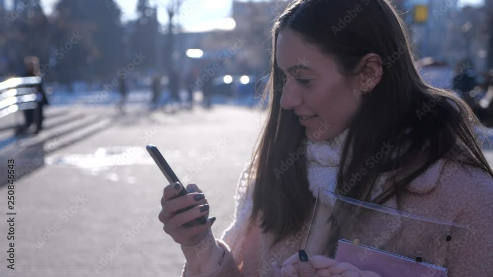 Portrait of smiling brunette woman in stylish coat and scarf, on city street with unsing smartphone