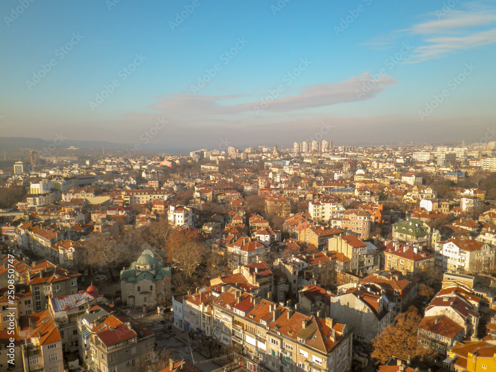 View from the height of the old town with low houses with tiled roofs and bare trees against the blue sky and gray clouds on the horizon