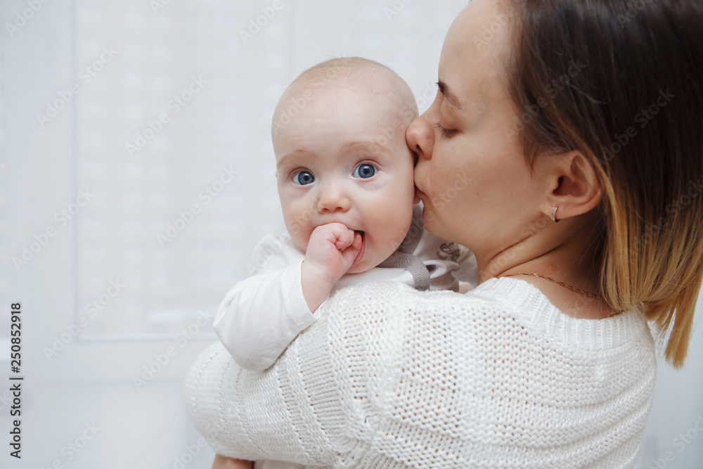 Young happy loving mother plays kisses her baby daughter in her arms against the window. Maternal care. Childcare. Close-up. Young happy loving mother plays kisses her baby daughter in her arms against the window. Maternal care. Childcare. Close-up.