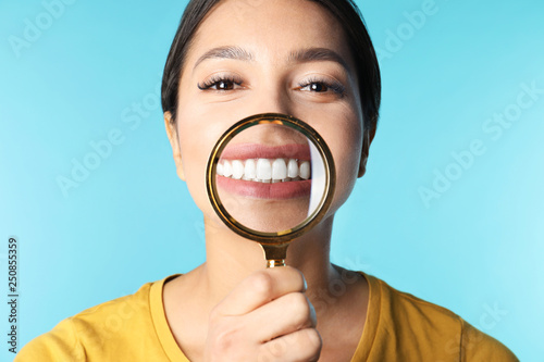 Young woman with healthy teeth and magnifier on color background, closeup