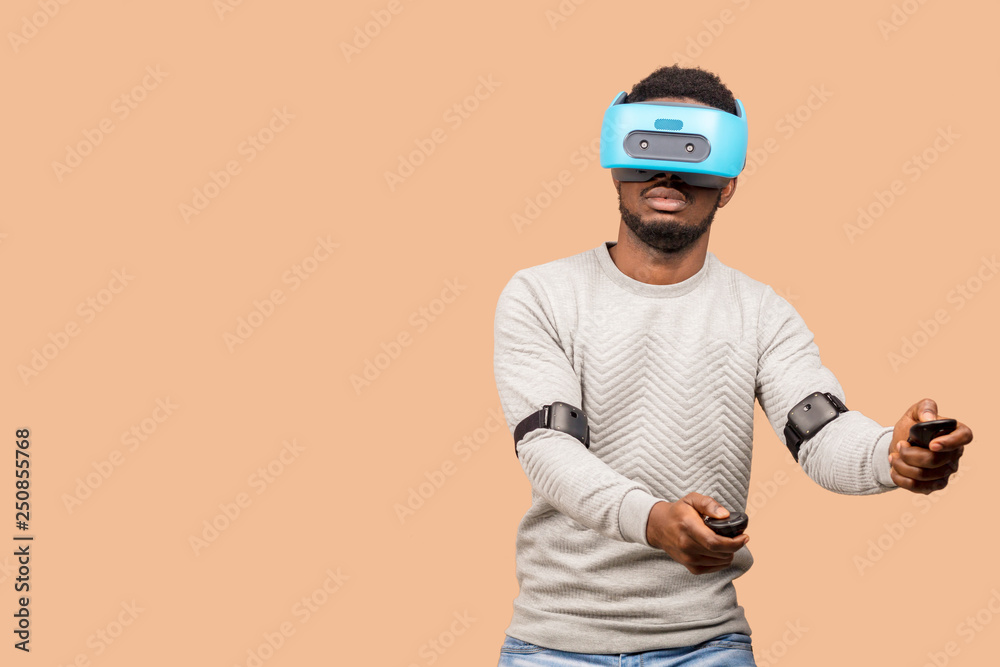 Studio shot of African american man using virtual reality headset, operating with joystick in hands, moving over beige background Studio shot of African american man using virtual reality headset, operating with joystick in hands, moving over beige background