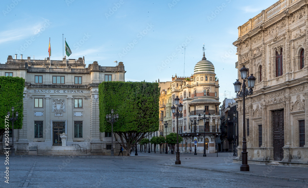 Fototapeta premium A look across the deserted cobbled Plaza de San Francisco towards the Banco de España & the lovely 'Confiteria Filella' on the 'Avenida de la Constitucion' in Seville just after sunrise.