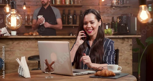 Pretty, brunette woman answers a phone call while working on her laptop remotly. Working as a freelancer from a coffee shop.