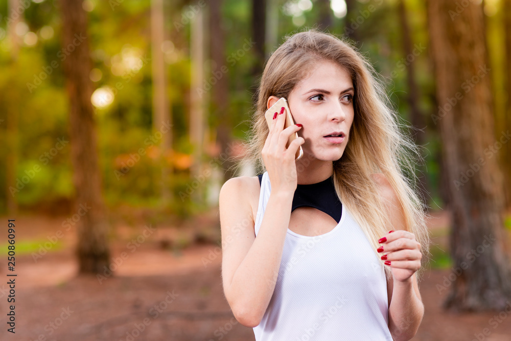 Beautiful blond young woman in park at sunset talking on mobile phone - Image.