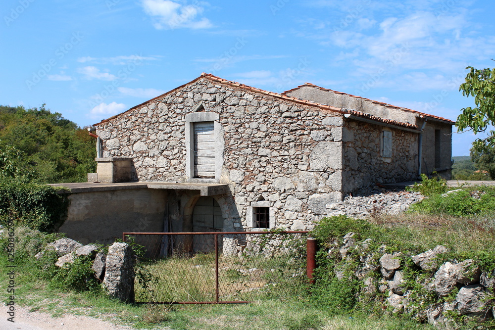 Abandoned family house built in traditional stone style with wooden window blinds and wooden doors surrounded with uncut grass and forest vegetation on cloudy blue sky background on warm sunny day Abandoned family house built in traditional stone style with wooden window blinds and wooden doors surrounded with uncut grass and forest vegetation on cloudy blue sky background on warm sunny day