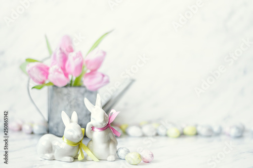 Easter table with porcelain bunnies  eggs and metal watering can with pink tulips on marble background.