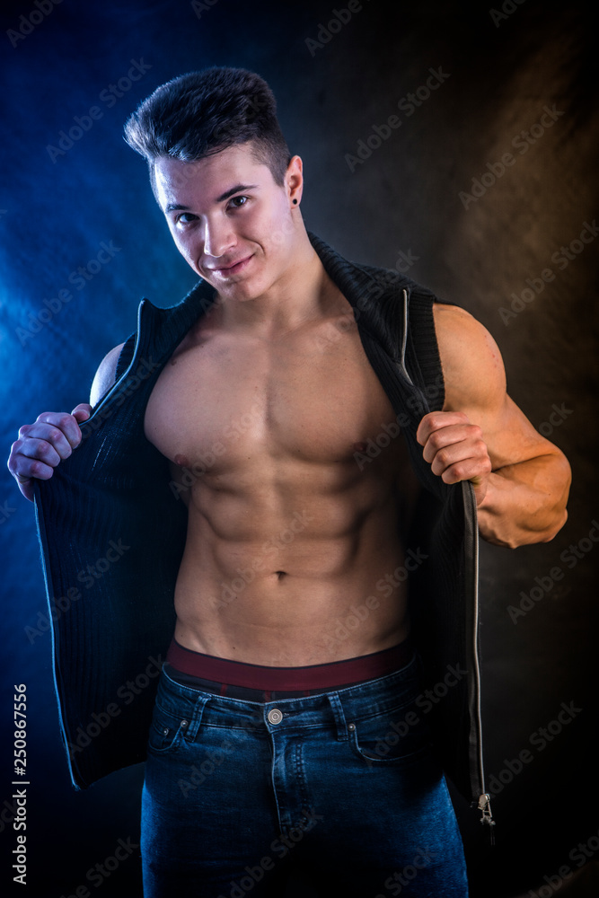 Handsome young muscular man shirtless wearing jeans and sleeveless vest, on black background in studio shot Handsome young muscular man shirtless wearing jeans and sleeveless vest, on black background in studio shot