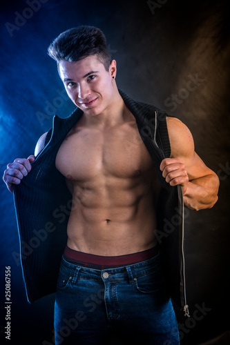 Handsome young muscular man shirtless wearing jeans and sleeveless vest, on black background in studio shot