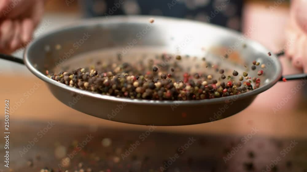 SLOW MOTION, MACRO, DOF: Unrecognizable woman toasting colorful peppercorns in a metal pan. Female chef mixing fragrant multicolored pepper seeds in a frying pan. Peppercorns flying around kitchen.