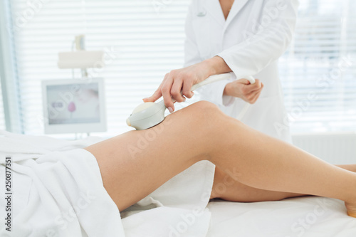 Unrecognizable female cosmetologist doing Rf lifting procedure on woman s long slim legs in a beauty parlor. Treatment of overweight and flabby skin.