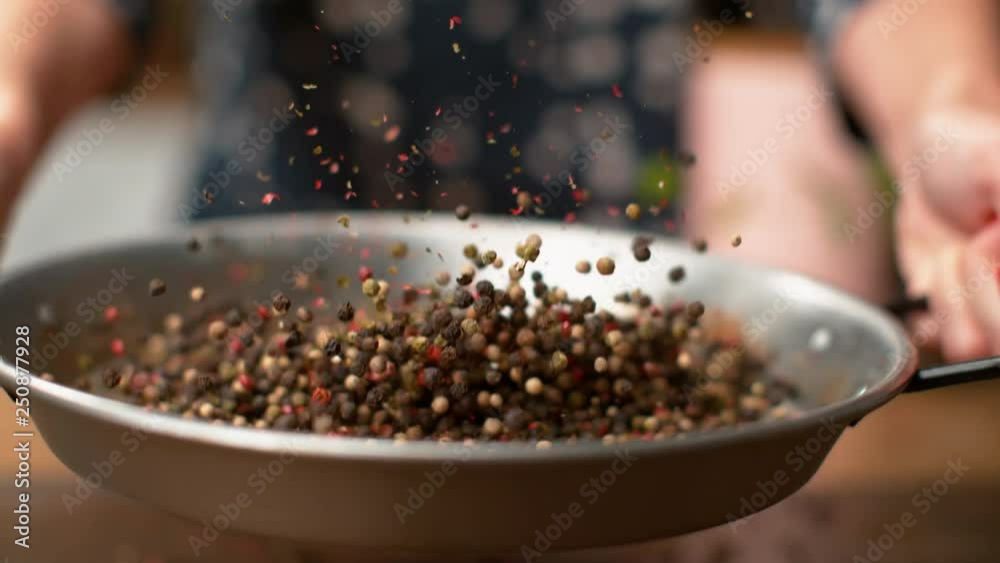MACRO, DOF, SLOW MOTION: Peppercorns flying around the kitchen as young woman roasts them in a hot metal pan. Unrecognizable female chef toasting aromatic and colorful peppercorns in a frying pan.