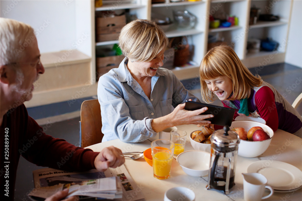 High angle view of grandparents and granddaughter sitting by dinning table, looking at digital tablet, laughing High angle view of grandparents and granddaughter sitting by dinning table, looking at digital tablet, laughing