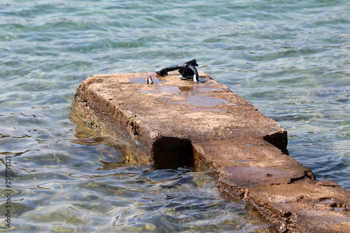 Improvised old small rocky concrete pier surrounded with clear calm sea with diving half mask and snorkel on warm sunny day