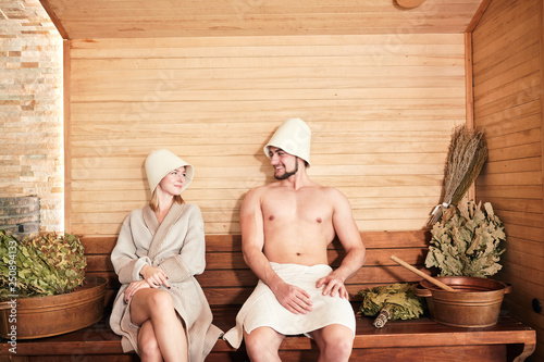 Quadro su tela Young couple of tourist relaxing in a sauna sitting on wooden beach with bath bowls and brooms