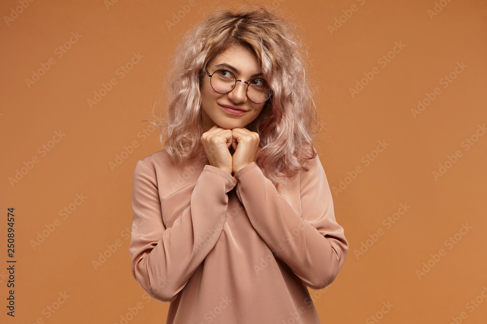 Adorable timid young woman wearing trendy round eyeglasses, holding clasped hands under her chin and looking away, having shy mysterious smile. Cuteness, youth, beauty, style and fashion concept