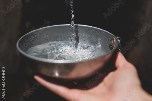 Hands collecting water from fountain with metal bowl.