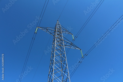 Mighty tall power line utility pole made of strong metal pipes with multiple electrical wires connected with glass insulators on clear blue sky background on warm sunny day