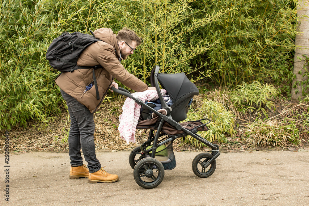 Obraz premium Bearded smiling father with a backpack and in yellow boots walking with a pram in a spring park shows care and love by covering a small child with a blanket.