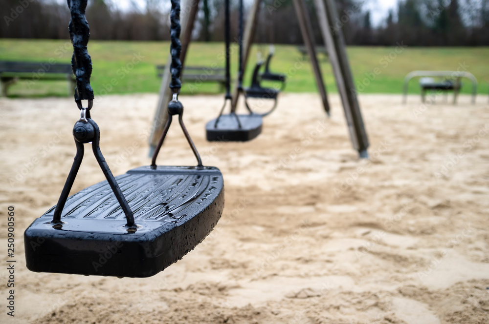 Wet swings after rain on a deserted playground, spring day, on a dim background of lawn and forest. - obrazy, fototapety, plakaty 