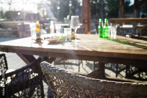 Dirty bottles, glasses, a wineglass and plates remained after eating and drinking drinks on a wooden table in the rays of the bright sun, in a street cafe.