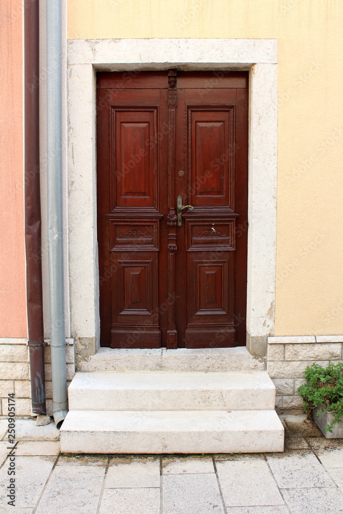 New hardwood family house entrance doors with decorative details and baroque style door handle mounted on stone frame wall next to two gutter pipes and stone steps in front on warm sunny day New hardwood family house entrance doors with decorative details and baroque style door handle mounted on stone frame wall next to two gutter pipes and stone steps in front on warm sunny day