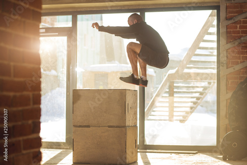 Box jump plyo exercise - young caucasian man doing functional workout at the well-lit gym in a sunny day, motion shot in touchdown phase. photo