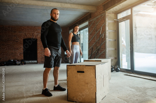 Handsome strong caucasian bearded man prepares to jumps onto a plyobox in a cross training gym with big panoramic windows and giant tire on background photo
