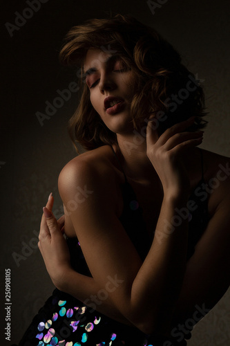 Seductive brunette model with curly hair wears sparkling dress posing with contrast light at studio
