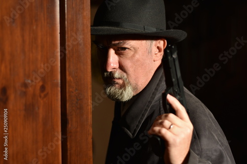 Slight three-quarter closeup of man in a fedora and raincoat (investigator or private detective) holding a gun near a wooden door. Interior scene selective focus