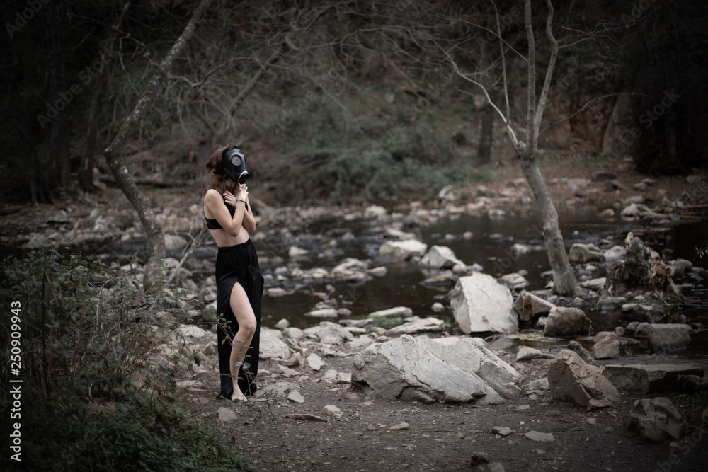 Obraz premium Side view of anonymous barefoot woman in black clothes and gas mask standing in amazing spooky forest